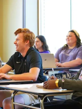 College students sitting at desks with laptops and notebooks, one raising their hand to participate.