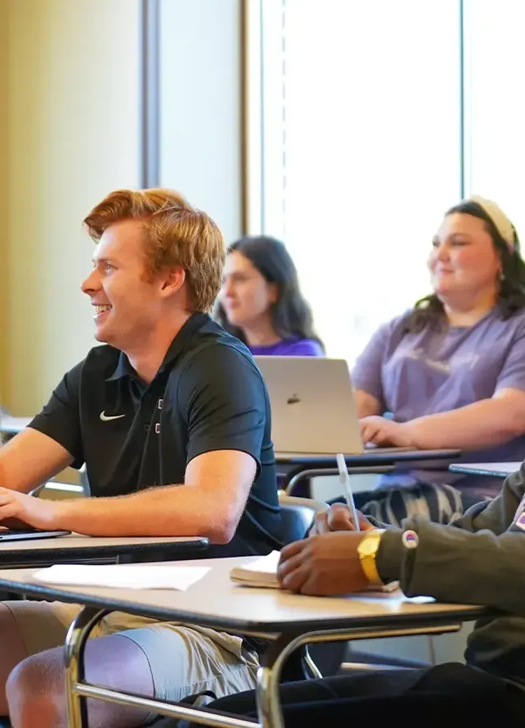 College students sitting at desks with laptops and notebooks, one raising their hand to participate.