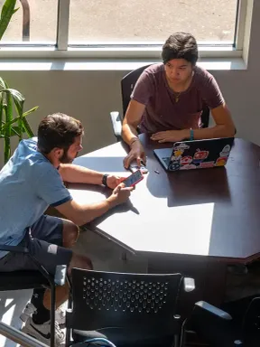 Two college students collaborating at a table with a laptop, seated near a sunlit window.