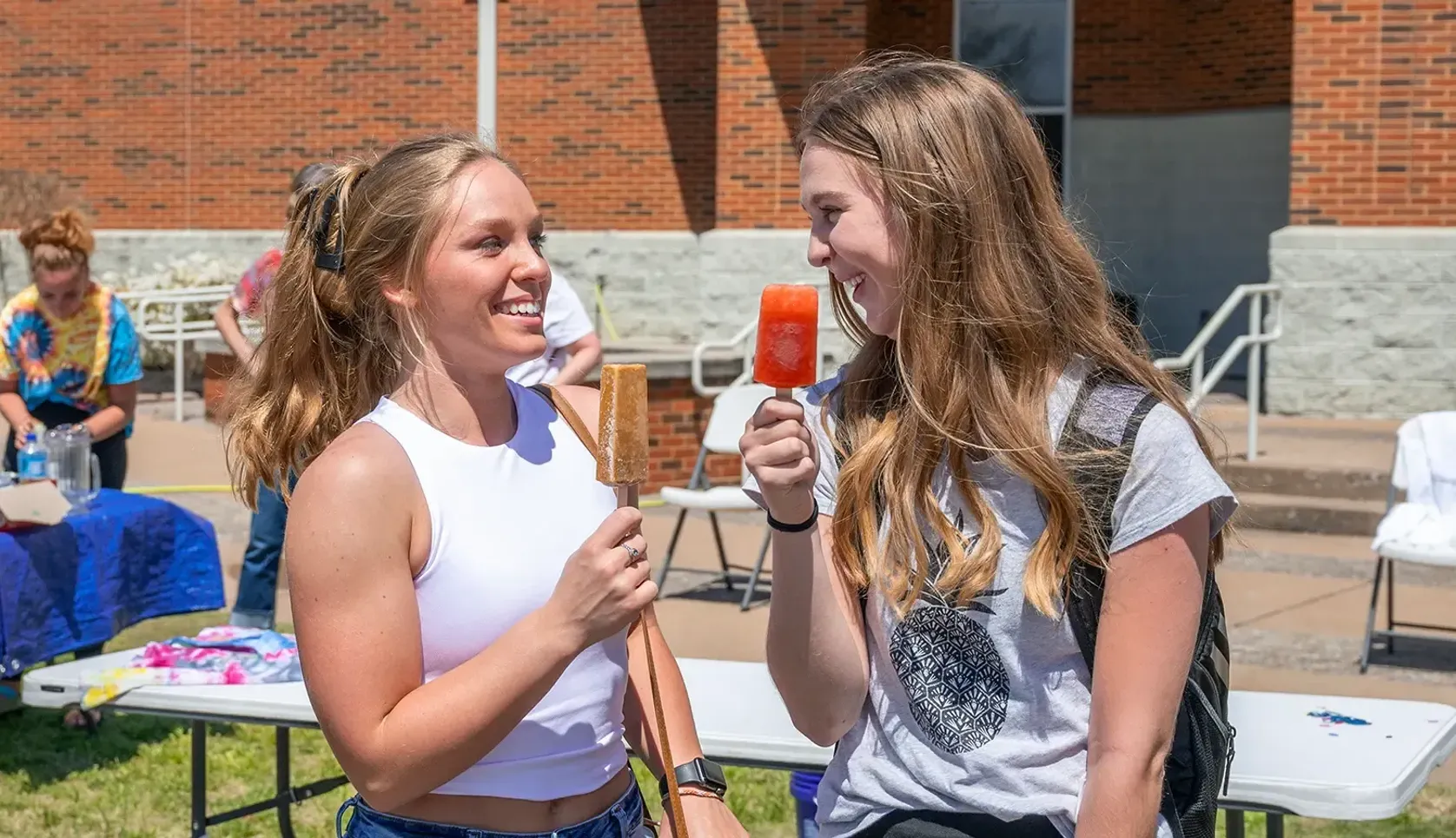 Two college students smiling and eating popsicles at an outdoor campus event on a sunny day.