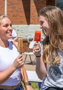 Two college students smiling and eating popsicles at an outdoor campus event on a sunny day.
