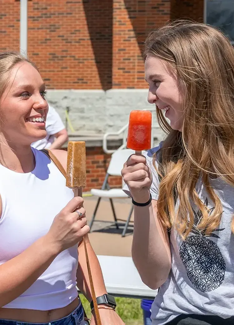 Two college students smiling and eating popsicles at an outdoor campus event on a sunny day.