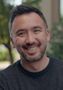 Lougan Bishop wearing a dark sweater, standing outdoors with blurred greenery and a building in the background.