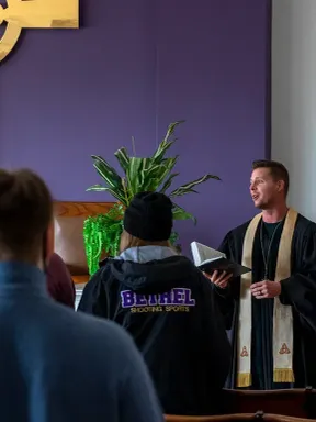 Students gather in a chapel as a robed minister leads a service beneath a large cross.