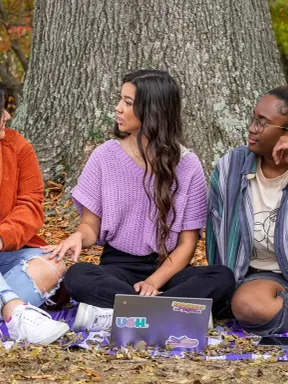 Three Bethel students sit together outdoors, talking and studying under a tree.