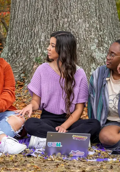 Three Bethel students sit together outdoors, talking and studying under a tree.