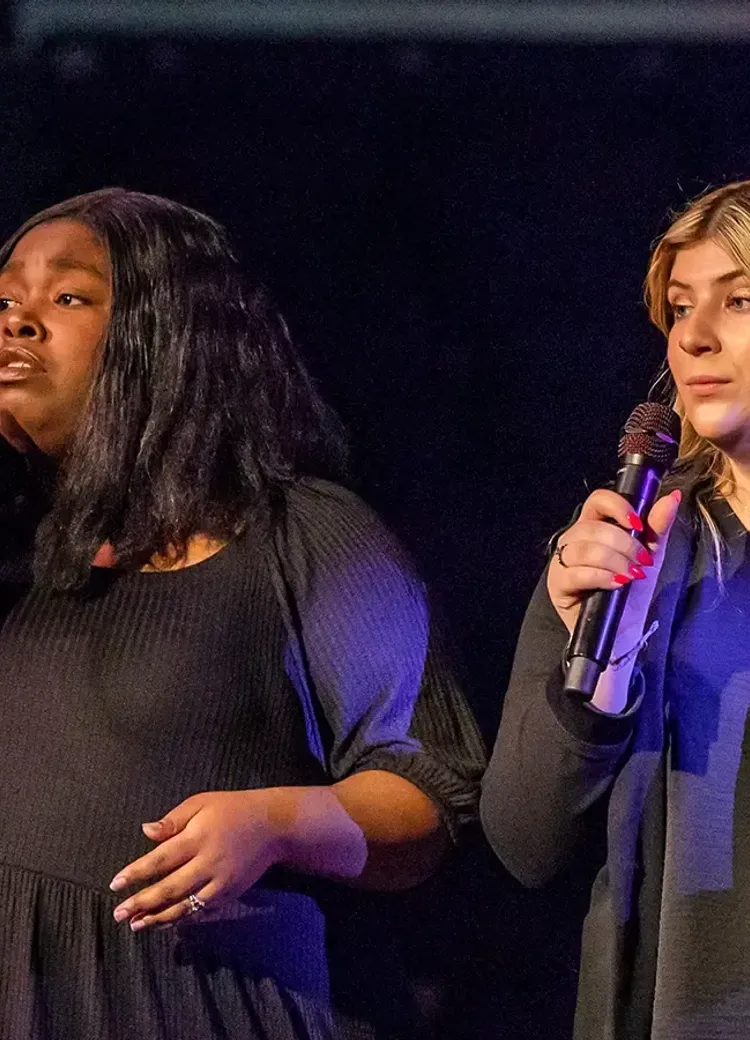 Two students sing into microphones during a performance on stage.