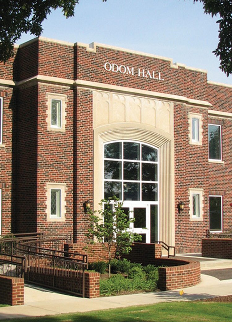 Exterior view of Odom Hall, a two-story red brick academic building featuring a large arched entryway with floor-to-ceiling windows, decorative stonework, and an accessible ramp leading to the front entrance, framed by trees and manicured landscaping.