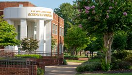Exterior view of the Ray and Linda Morris Science Complex, featuring a distinctive curved white facade with gold lettering, white columns, and an adjacent red brick wing, surrounded by lush landscaping, flowering crape myrtle trees, and a campus walkway on a sunny day.