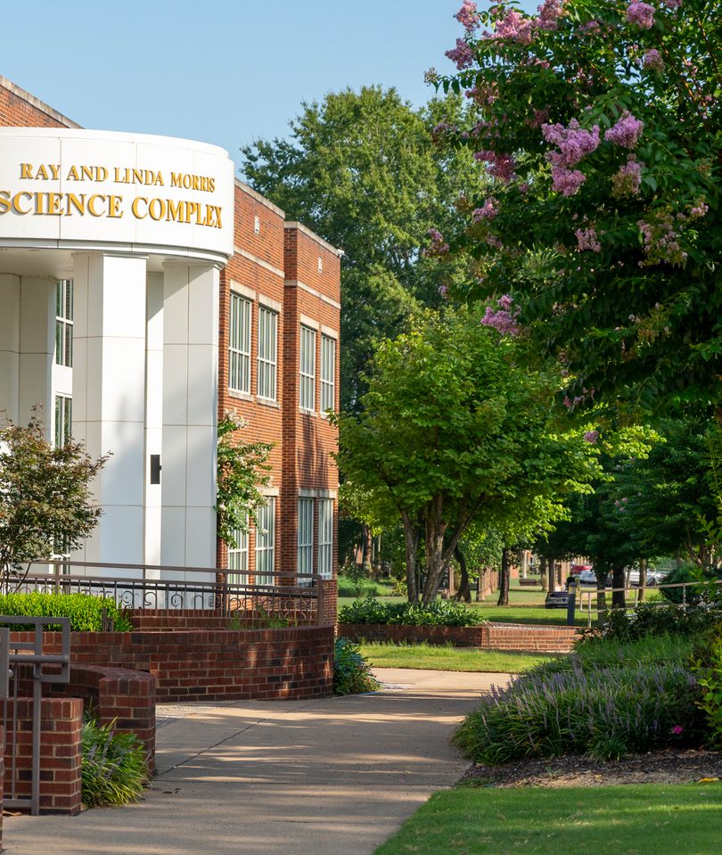 Exterior view of the Ray and Linda Morris Science Complex, featuring a distinctive curved white facade with gold lettering, white columns, and an adjacent red brick wing, surrounded by lush landscaping, flowering crape myrtle trees, and a campus walkway on a sunny day.